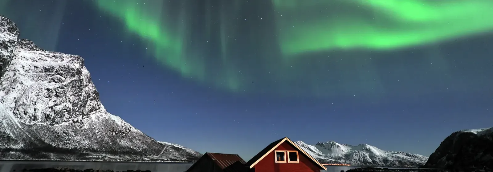 Ein rotes Haus bei Nacht mit Nordlichtern über schneebedeckten Bergen und klarem Himmel in Tromsö.