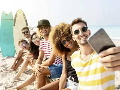 Eine Gruppe junger Menschen sitzt in eine Reihe am Strand und macht ein Selfie.