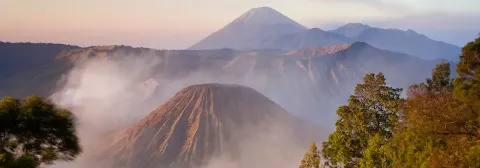 Vulkan Mount Bromo mit mehreren Kratern am Morgen eingehüllt von Nebelschwaden im Vordergrund.