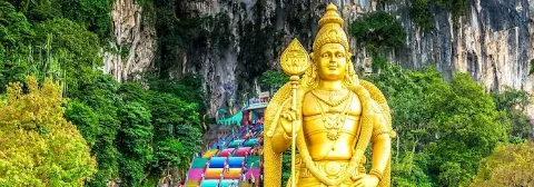 Eine goldene Statue von Lord Murugan vor bunten Treppen und grüner Natur vor den Batu-Höhlen.