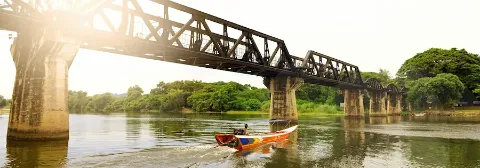 Ein Mann auf einem Langboot fährt auf dem Fluss Kwai in Thailand entlang einer Brücke mit grünem Ufer.