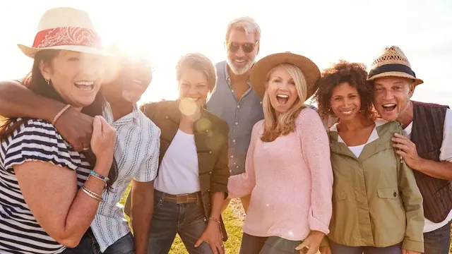 Gruppe reifer Freunde steht lachend eng beieinander im Gegenlicht der tiefstehenden Sonne, einige tragen Hüte.