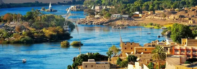 Panoramablick auf den Nil mit Booten, umgeben von üppiger Vegetation und Häusern der Stadt Assuan in Ägypten.
