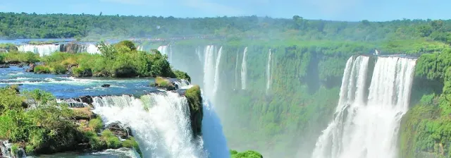 Die Iguazu Wasserfälle stürzen über grüne Felsen in eine Schlucht mit grünen Wäldern im Hintergrund und blauem Himmel.