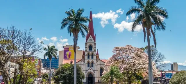 Frontalansicht der Kirche Iglesia de Nuestra Señora de la Merced im Stadtpark von San José, umgeben von Bäumen und Palmen bei sonnigem Wetter mit blauem Himmel.