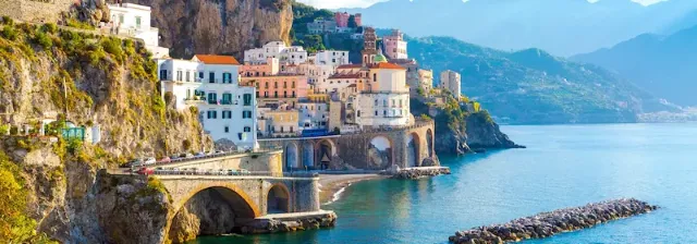 Küstenort Amalfi mit dicht gebauten Häusern auf Felsen, mehreren Brückenbögen und ruhigem Meer im Vordergrund.