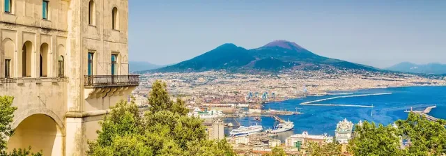 Panoramaaufnahme mit Blick über die Stadt Neapel mit Küste, Gebäuden und dem Vulkan Vesuv im Hintergrund.
