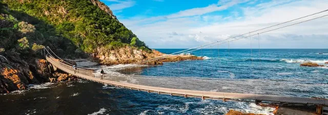Hängende Brücke über das Meer auf der Gartenroute im Tsitsikamma Nationalpark mit Küstenlandschaft im Hintergrund.