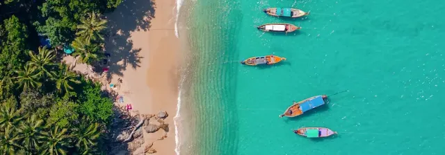 Luftaufnahme von vier Langbooten, die auf türkisfarbenem Meer nahe dem von Palmen gesäumten Sandstrandes treiben.