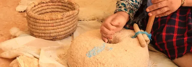 Berberfrau dreht Holzstab in rundem Mahlstein neben geflochtenem Korb als Vorbereitung zum Brot backen.