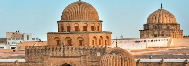 Panoramablick über die Stadt Kairouan mit zwei große Kuppelbauten mit Rundbögen und Mauern im Vordergrund.