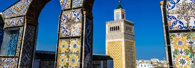 Verzierte Fliesenbögen der Großen Moschee Zitouna in Tunis mit einem Minarett im Hintergrund unter klarem Himmel.