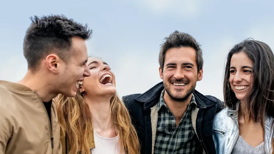 Vier Menschen stehen nebeneinander und lachen in Nahaufnahme vor blauem Himmel.