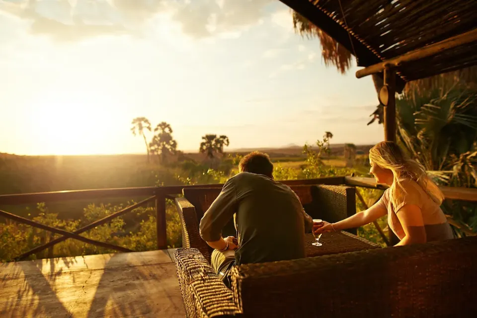 Zwei Reisegäste stoßen auf der Terrasse einer Lodge in Afrika bei Sonnenuntergang an