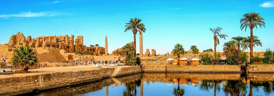 Panorama der antiken Ruinen von Luxor mit Palmen und einem ruhigen Wasserlauf.