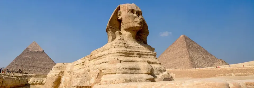Die Große Sphinx von Gizeh im Vordergrund mit zwei Pyramiden im Hintergrund unter blauem Himmel.