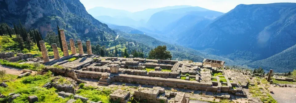 Ruine des Apollontempel in Delphi mit mehreren Säulen und rechteckigen Grundmauern vor einer Gebirgslandschaft.