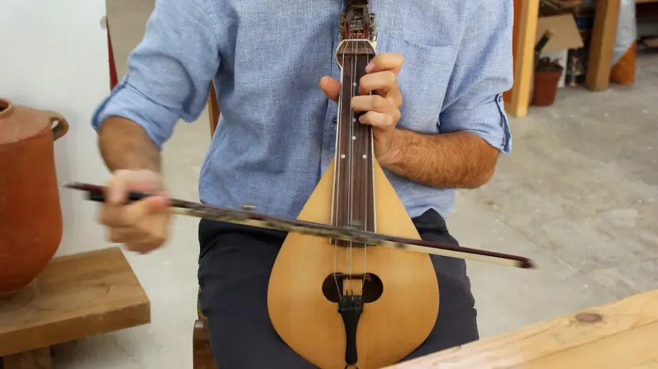 Ein Musiker spielt eine Rebetiko mit einem Bogen in einer Werkstatt, umgeben von Töpfen und Holz.