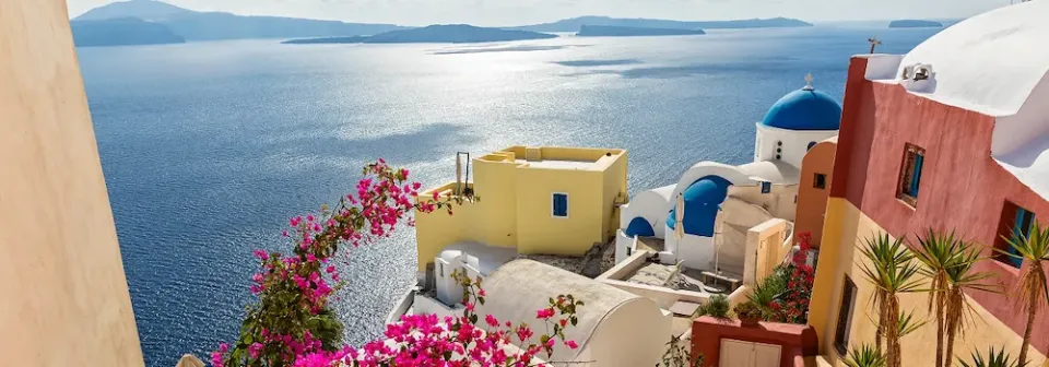 Blick auf weiße Häuser mit blauen Kuppeln und rosa Bougainvillea auf der Insel Santorin mit Küste mit ruhigem Meer.