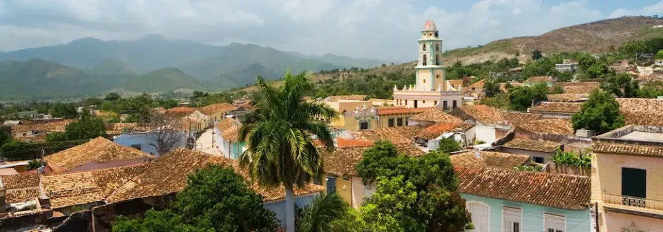 Blick auf die Stadt Trinidad auf Kuba mit roten Ziegeldächern, Palmen und einem Kirchturm vor einer hügeligen Landschaft.