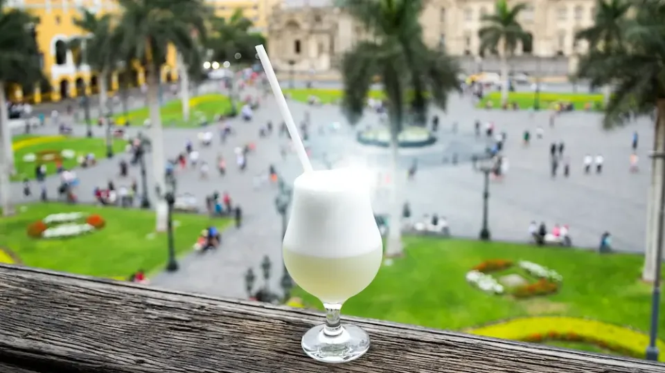 Cocktail Pisco Sour in einem Glas mit Strohhalm steht auf einem Balkon vor dem belebten Hauptplatz in Lima.