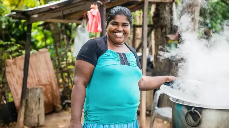 Einheimische Frau mit Zopf in Sri Lanka und türkisfarbenem Kleid steht neben großem dampfendem Topf.


