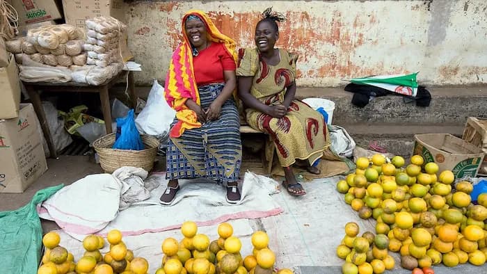 Zwei afrikanische Frauen in gemusterten Kleidern sitzen neben einem Marktstand mit Orangen und anderen Früchten.