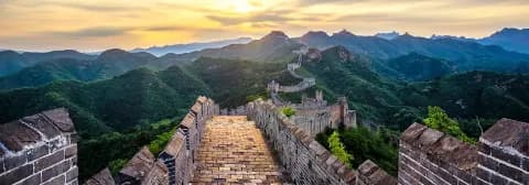 Panorama über die Chinesische Mauer bei Sonnenuntergang, umgeben von grünen Hügeln.
