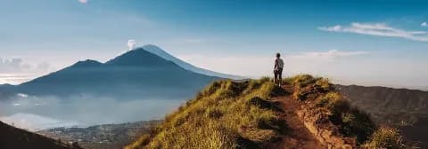 Person steht auf einem grasbewachsenen Bergrücken mit Blick auf den Vulkan Batur und einen See.