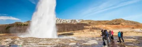 Touristen beobachten einen Geysir in Island, der eine weiße Wasserfontäne sprudelt, umgeben von einer Landschaft mit Bergen.