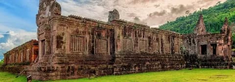 Hinduistischer Tempel Wat Phou in Laos steht auf einer grünen Wiese mit Bäumen im Hintergrund.