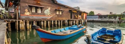 Blick auf ein traditionelles Holzhaus am Wasser mit bunten Booten in George Town auf der Insel Penang.