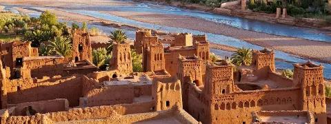 Stadt Ait Ben Haddou aus Lehmziegelbauten mit Zinnen und Türmen vor einem breiten Flussbett und Palmen im Hintergrund.