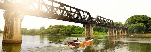 Ein Mann auf einem Langboot fährt auf dem Fluss Kwai in Thailand entlang einer Brücke mit grünem Ufer.