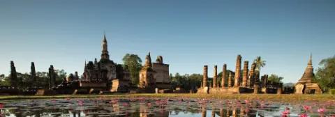 Panoramablick auf die Tempelruinen von Sukhothai in Thailand mit einem Seerosenteich unter klarem Himmel.