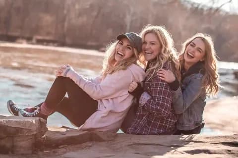 Drei lachende junge Frauen sitzen nebeneinander am Wasser.
