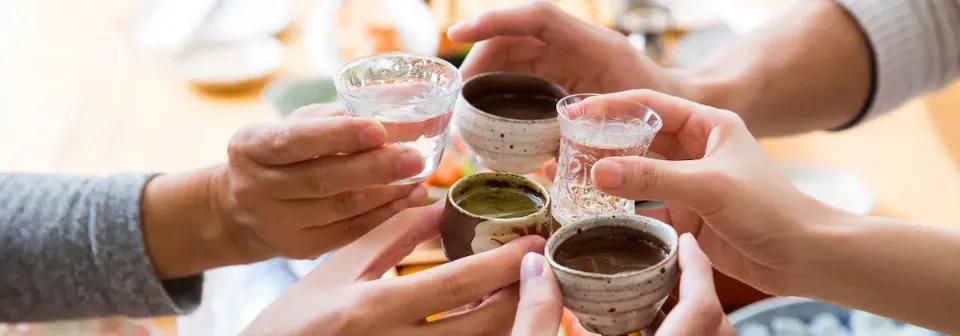 Menschliche Hände stoßen mit kleinen Gläsern, gefüllt mit Sake, an.