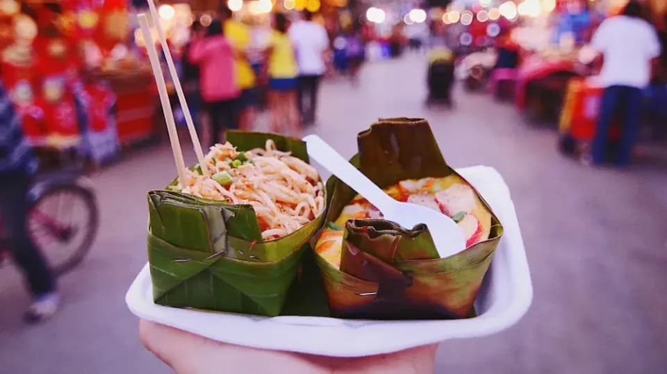Hand hält Straßenessen Nudeln und Gemüse in Bananenblättern auf einem Styroportablett auf einem Nachtmarkt.
