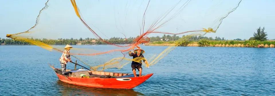Zwei Vietnamesen stehen auf einem Boot und sie werfen ein Fischernetz in den Fluss aus.
