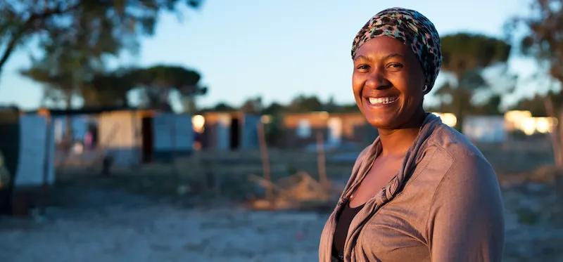 Eine afrikanische Frau lächelt im frühen Morgenlicht, umgeben von den Häusern ihrer Gemeinde.