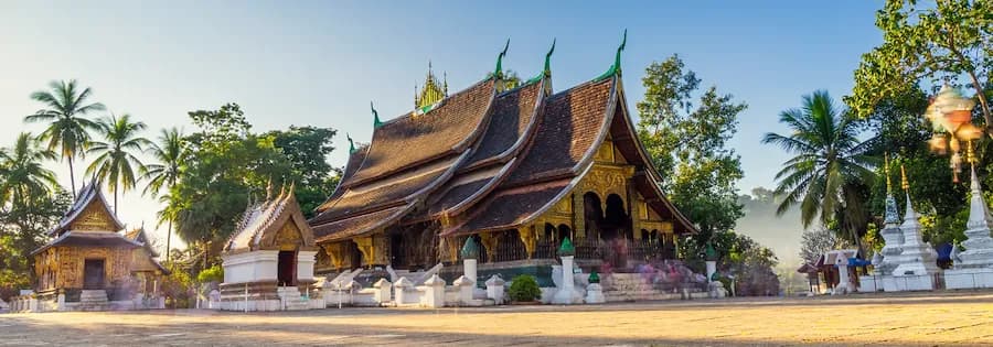 Tempel in Luang Prabang mit geschwungenem Dach und reich verzierten Fassaden zwischen Palmen und Stupas.