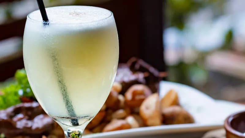Glas mit Cocktail Pisco Sour und einem schwarzen Strohhalm mit Essen im Hintergrund.