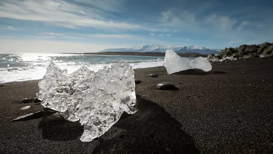 Glitzernde Eisbrocken am schwarzen Lavastrand Diamond Beach in Island, mit Meer und Bergen im Hintergrund.