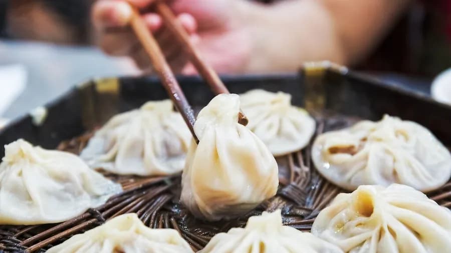 Hand hält mit Stäbchen eine chinesische Teigtasche auf einem bambusähnlichen Korb mit weiteren Teigtaschen.