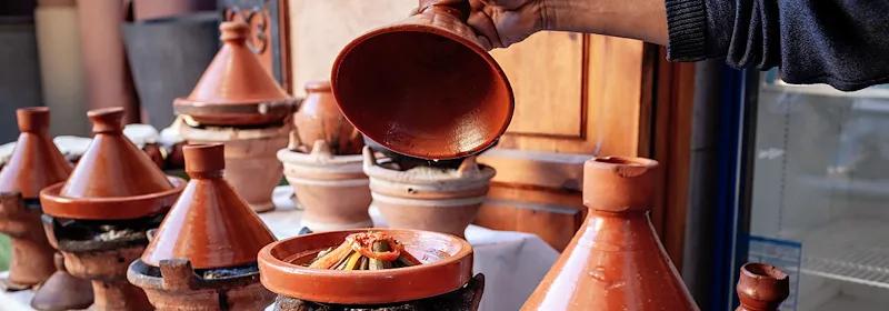 Männerhand hebt den Deckel eines Tajine in der Medina von Marrakesch und es erscheint eine Gemüse-Tajine.