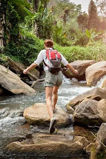 Individuelle Reisende mit Rucksack springt über Steine eines Flusses im Wald