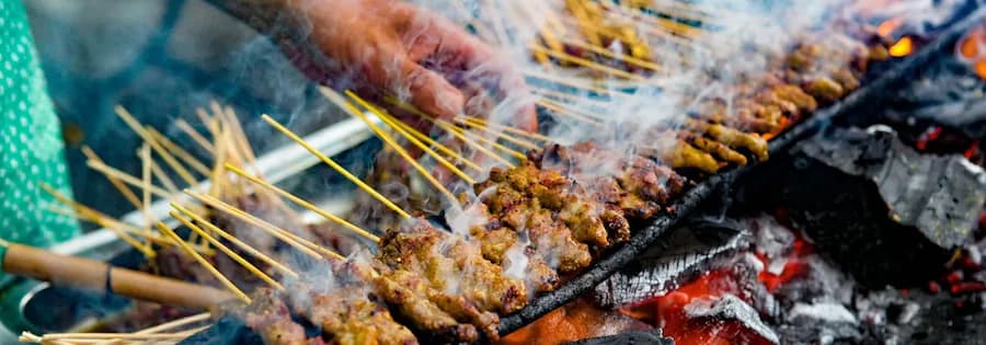 Hand dreht mehrere Satay-Fleischspieße auf einem Grill über offener Glut mit aufsteigendem Rauch.