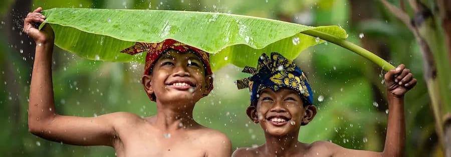 Zwei fröhliche Kinder halten ein großes Bananenblatt über ihrem Kopf, während Regen auf sie fällt.