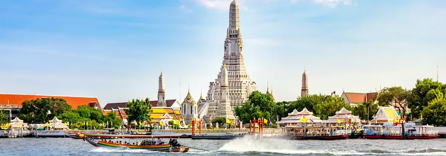 Boot auf dem Fluss mit Tempel Wat Arun in Bangkok am Ufer und Gebäuden im Hintergrund unter klarem Himmel.