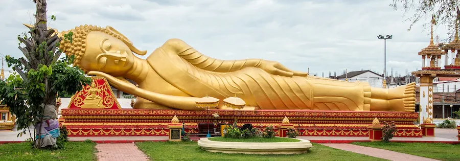 Große goldene liegende Buddha-Statue Pha That Luang auf verziertem Sockel unter bewölktem Himmel.